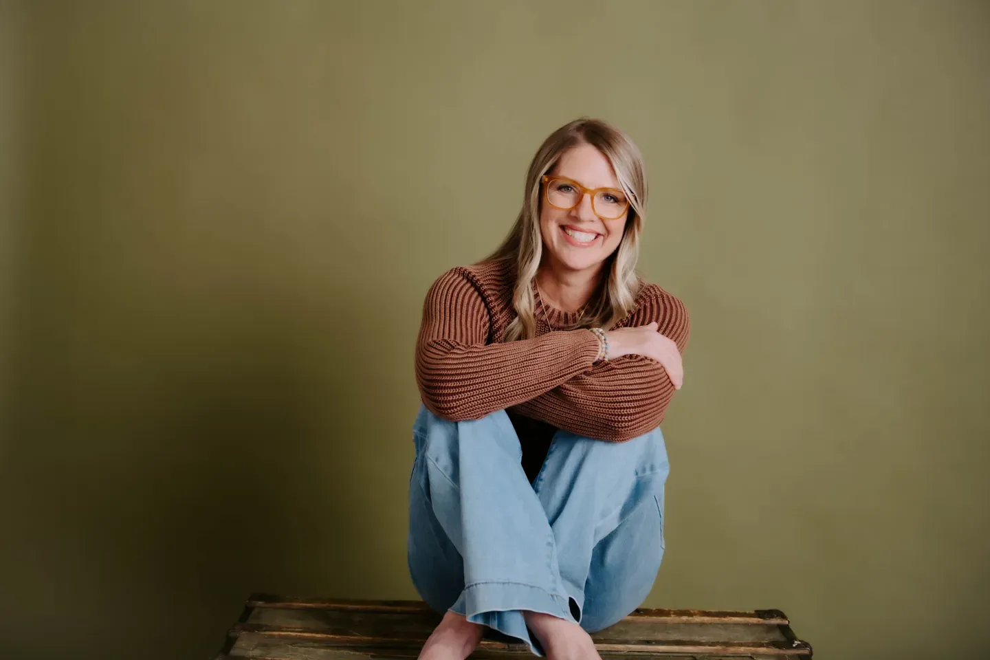Julie Holly smiling with her arms around her knees against an olive wall