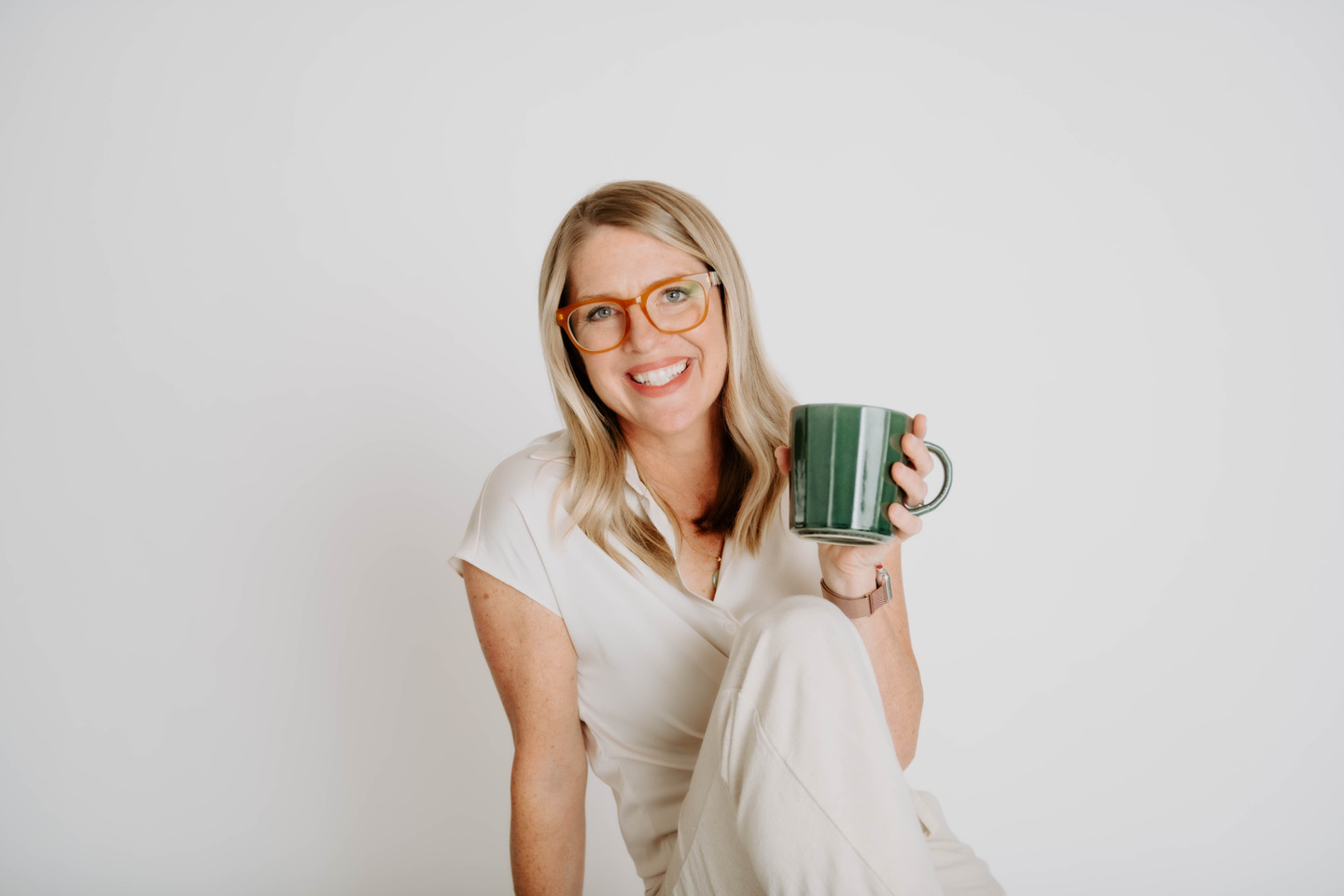 Julie Holly smiling in amber glasses and a cream outfit, holding a green mug.