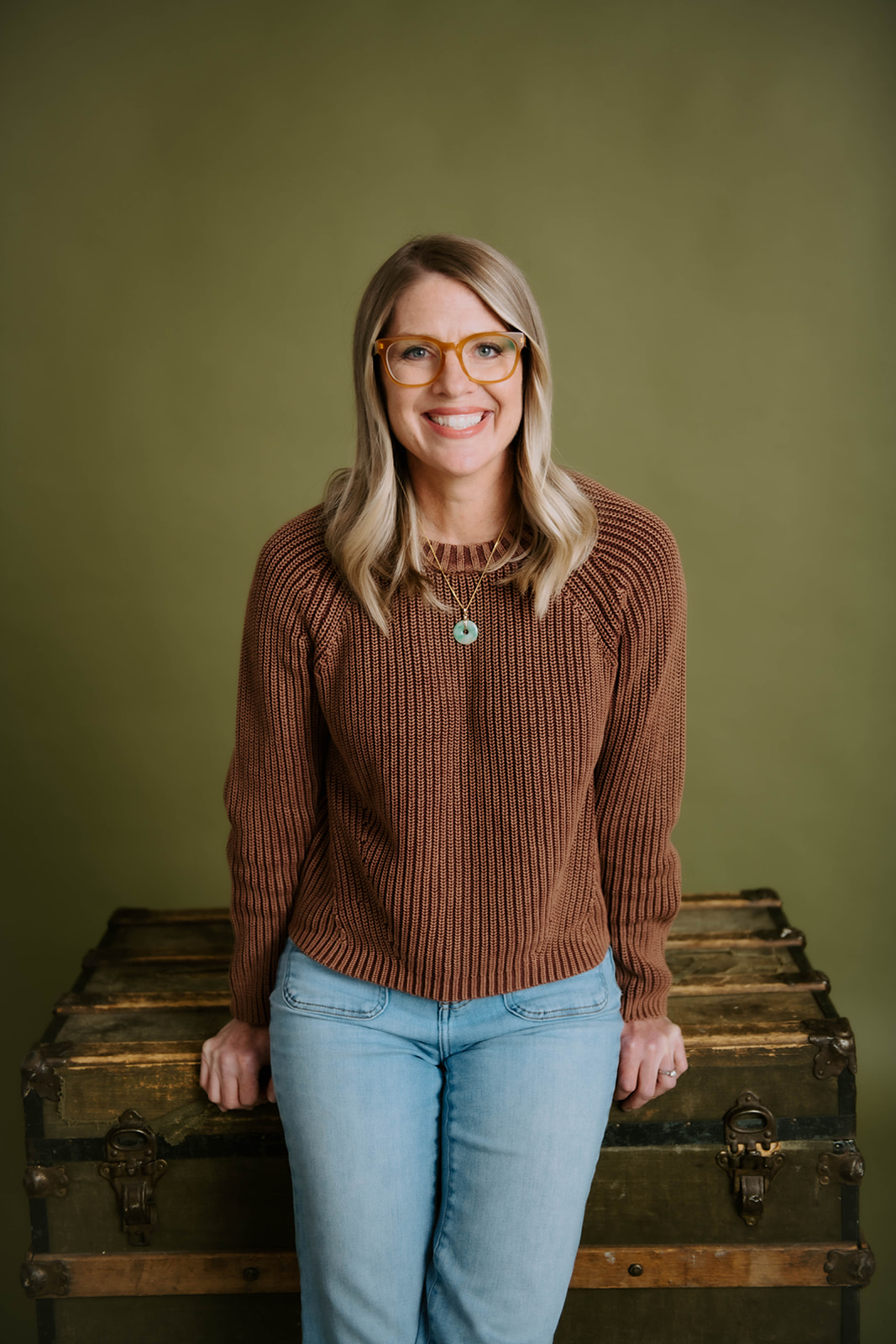Julie Holly standing in amber glasses, a brown sweater, and jeans beside a wooden trunk