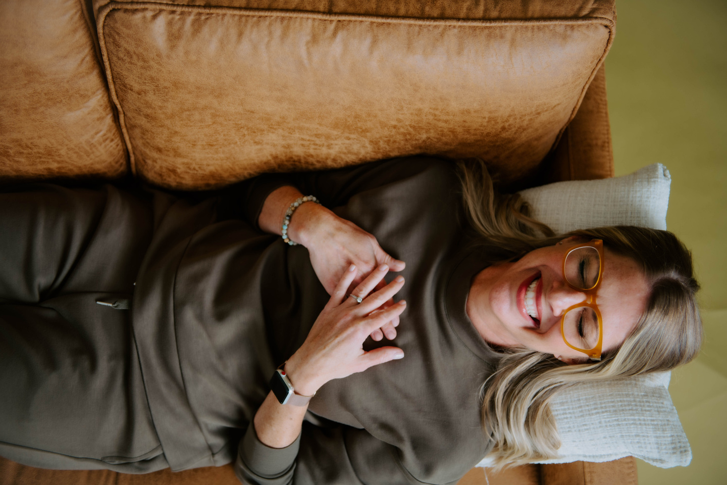 Julie Holly laughing while reclining on a leather sofa, with her hands resting over her heart.